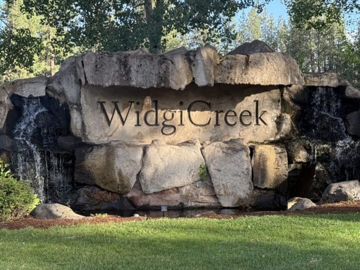 A large stone sign reading Widgi Creek is set into a rock formation with small waterfalls on both sides. Green grass and trees surround this Bend Oregon Golf Course landmark, as sunlight casts shadows across the rocks.