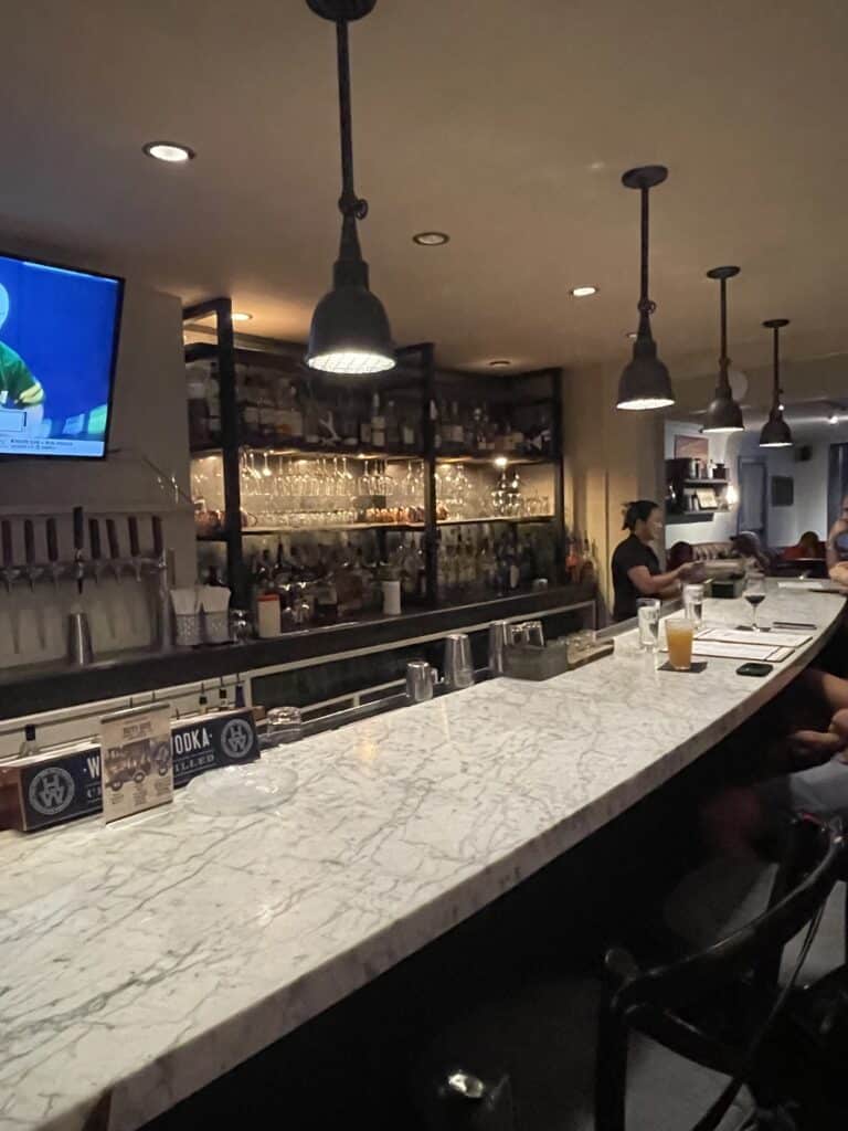 Vintage Restaurant Bar a Marble Topped Bar with Hanging Pendant Lights Shelves Stocked with Liquor Bottles and Glassware Behind It in New Market Md a Tv Shows a Sports Event As Several People Sit at the Bar One with a Drink in a Dimly Lit Cozy Atmosphere