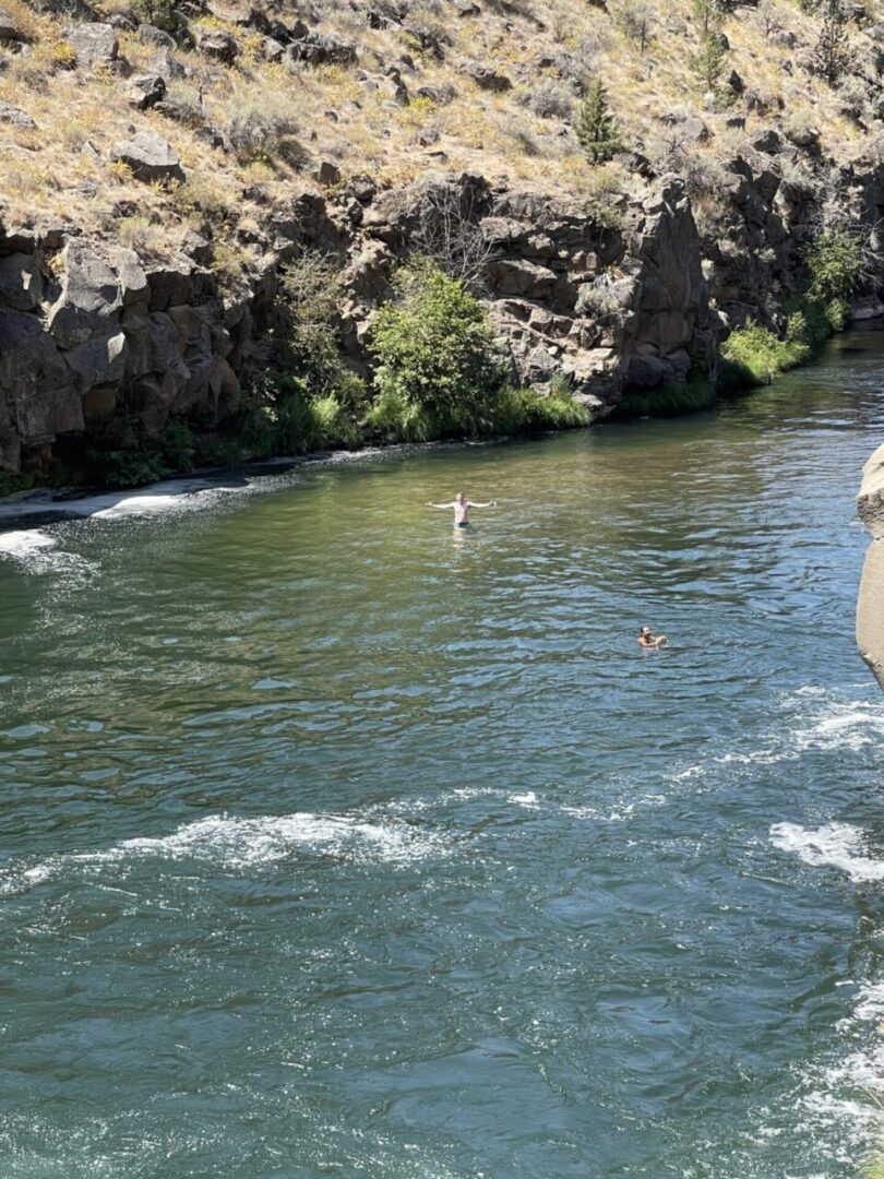 Deschutes River After Steelhead Falls a River with Rippling Blue Green Water Flows Past Rocky Shrub Covered Banks Near Steelhead Falls Two People Are Swimming One in the Center Raising Their Arms As Sunlight Illuminates the Water and Dry Terrain Above the Rocks