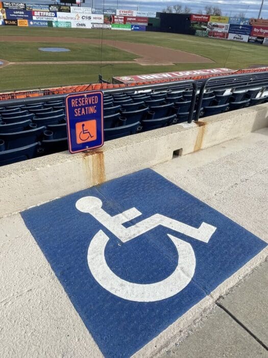 a Large Blue and White Wheelchair Accessible Symbol is Painted on Concrete Near Stadium Seating Above It a Sign Reads Reserved Seating in the Background an Empty Baseball Field and Frederick Keys Schedule Advertisement Boards Are Visible