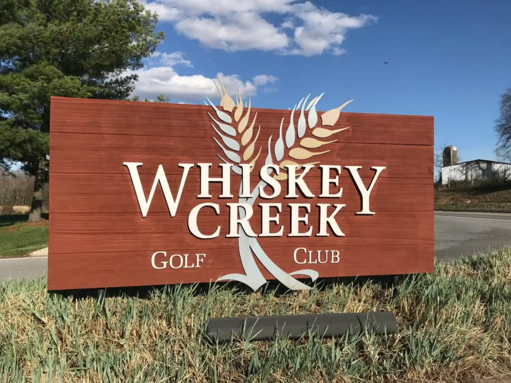 Whiskey Creek Golf Club Sign Scaled a Large Wooden Sign Reading Whiskey Creek Golf Club in White Letters with Two Stylized Wheat Stalks Crossing Behind the Text Stands on Grass Beside a Road in New Market Md Trees a Blue Sky with Clouds and Distant Farm Buildings Are in the Background