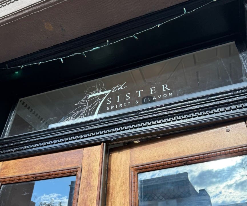 a Close up of a Glass Transom Above Wooden Double Doors the Glass Reads 7th Sister Spirit  Flavorone of the Best Bars in Frederickwith an Abstract Floral Line Drawing Beside the Text White String Lights and Blue Sky Reflect in the Glass