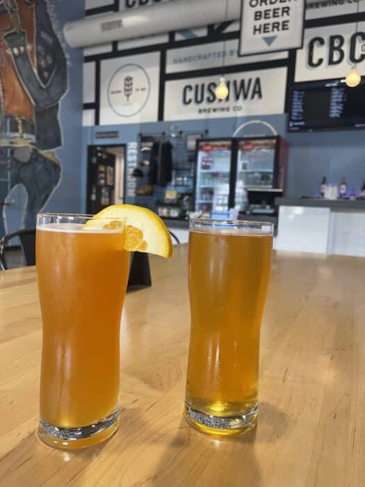 Two Tall Glasses of Beer Sit on a Light Wooden Table at One of the Best Breweries in Frederick Md One is Hazy Orange with an Orange Slice the Other Clear Golden Behind Them is a Taproom with a Large Menu Board and Signs Reading Custowa