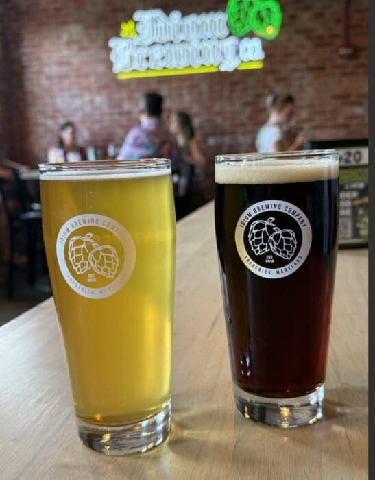 Two Pint Glasses of Beer Sit on a Wooden Table at One of the Best Breweries in Frederick Md One Beer is Pale Yellow and Hazy the Other Dark Brown Both Feature a Twin Brewing Company Logo with a Blurred Brewery Sign and People in the Background