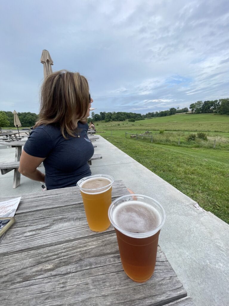 Nicole at Freys Brewery Mount Airy a Woman with Shoulder Length Hair Sits at a Wooden Picnic Table Outdoors in New Market Md Gazing Toward a Green Field Under a Cloudy Sky Two Plastic Cups of Beer Rest on the Table with Umbrellas and More Tables Visible in the Background