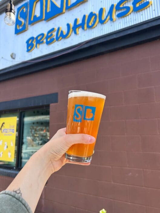 a Hand Holds a Pint of Hazy Amber Beer with a Blue Sb Logo in Front of a Brown Building a Sign Above Reads Sand Brewhouse in Bold Yellow and Blue Lettersone of the Best Breweries in Frederick Md a Tattoo is Visible on the Persons Wrist