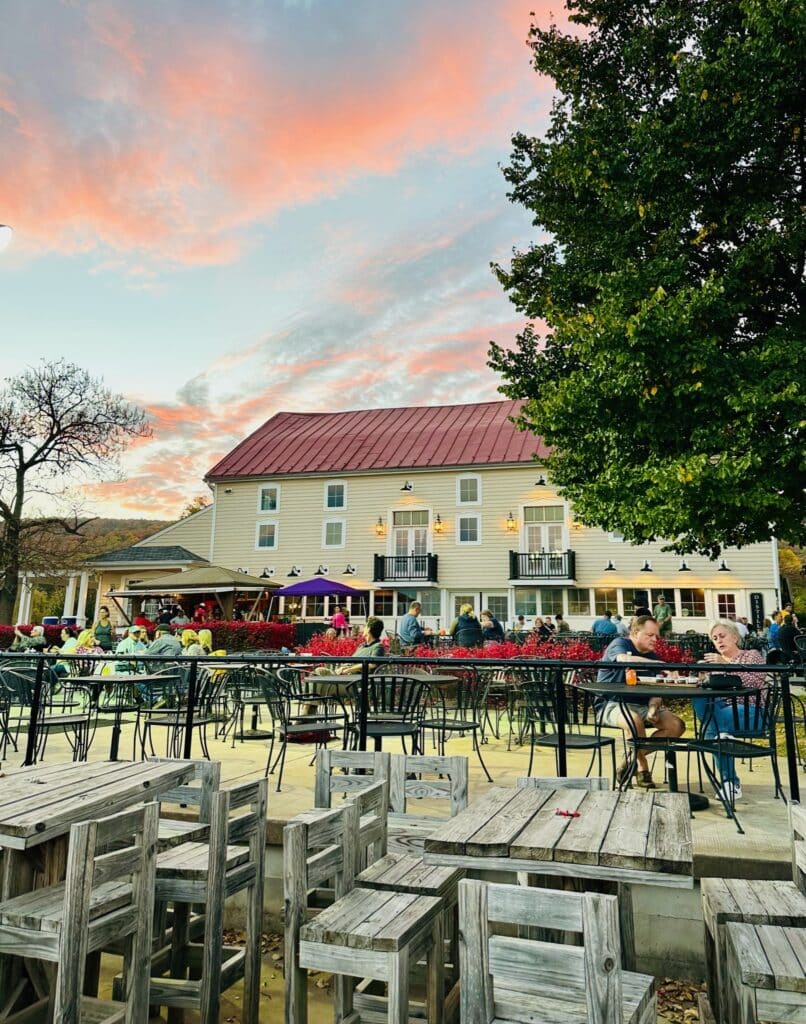 An outdoor dining area with wooden and metal tables and chairs is filled with people socializing. Behind them stands a large beige building, similar to some hotels in Frederick MD, under a colorful sunset sky framed by green trees.