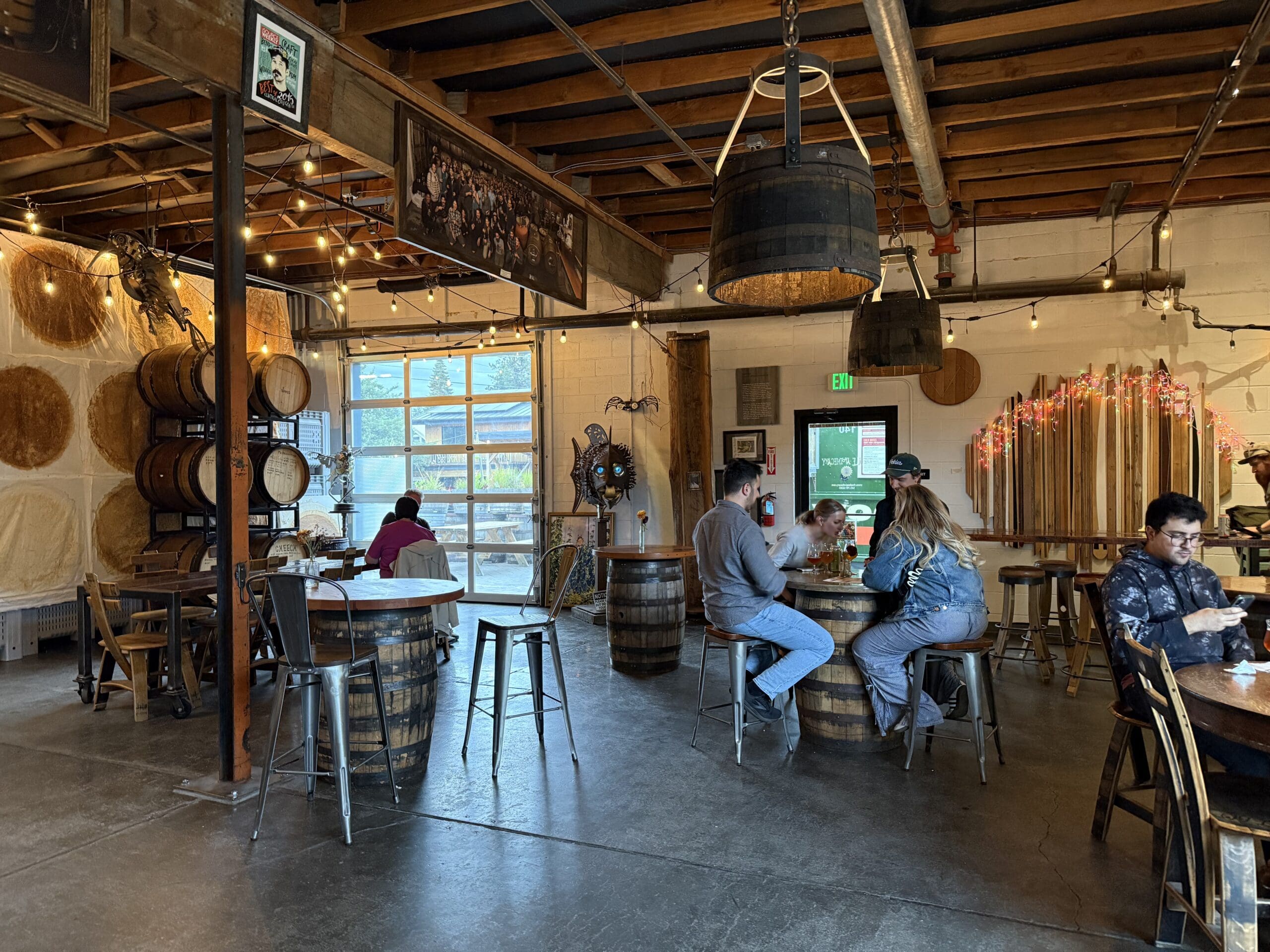 Industrial Style Brewery Taproom with Exposed Beams Hanging Lights and Stacked Barrels on the Left Wall Like Many Breweries in Bend Oregon People Sit at Round Barrel Tables Chatting and Drinking in a Cozy Welcoming Atmosphere Filled with Natural Light