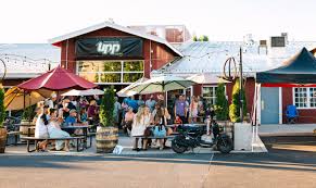 a Lively Outdoor Patio at One of the Top Breweries in Bend Oregon with People Sitting at Picnic Tables Under Colorful Umbrellas Socializing the Red Brick Building Has a Sign That Reads Lpp String Lights Glow Above and a Scooter is Parked in Front
