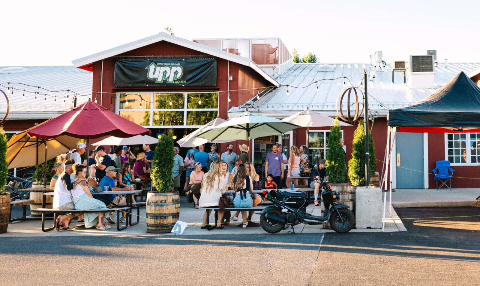 a Lively Group Gathers at Outdoor Picnic Tables Under Umbrellas Outside Tapp House One of the Popular Bend Breweries String Lights Barrel Planters and a Black Scooter Add to the Welcoming Vibe in Front of the Red Building