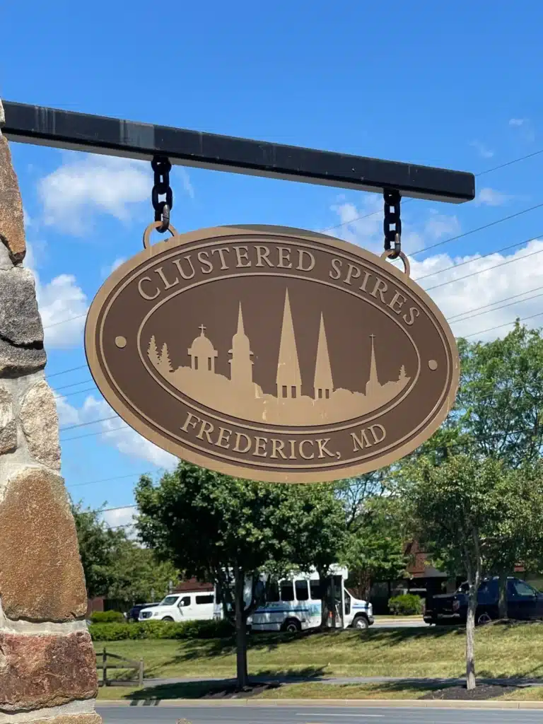 Clustered Spires Frederick Md Scaled Oval Tan Sign Reading Clustered Spires Frederick Md Featuring Church Spires Silhouettes Hangs from a Metal Bracket on a Stone Pillar in the Background Are Trees Parked White Vans and a Blue Sky with Clouds Near New Market Md