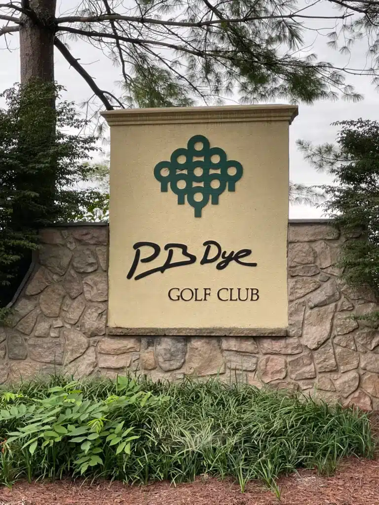 Pb Dye Golf Course Scaled a Stone Wall Displays a Beige Sign with a Green Abstract Golf Ball Logo Above the Words Pb Dye Golf Club in Black Script Bushes Tall Grass and Trees Surround the Sign Near New Market Md the Sky is Overcast