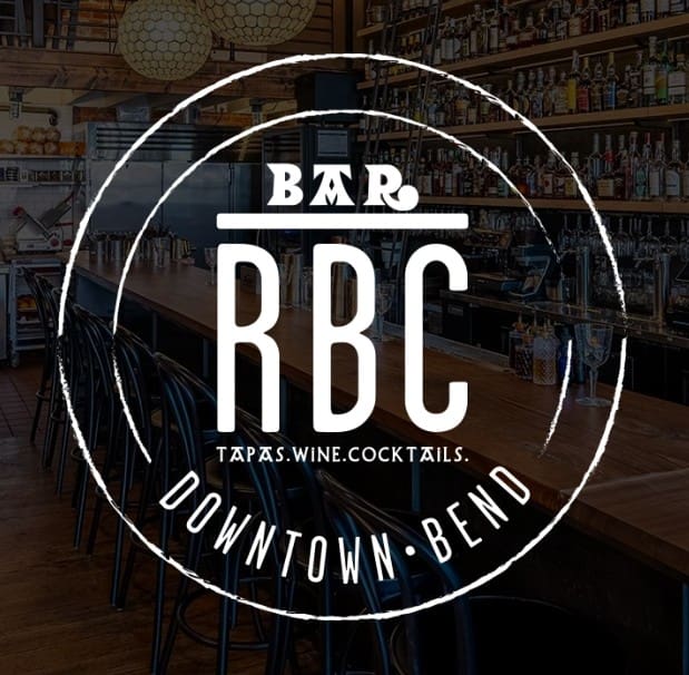 a Modern Bar with a Long Wooden Counter and Black Stools Shelves of Bottles Behind Overlaid Text Reads Bar Rbc Tapaswinecocktails Downtown Bendone of the Standout Bend Oregon Restaurantshighlighted in Bold White Letters Within a Circular Frame