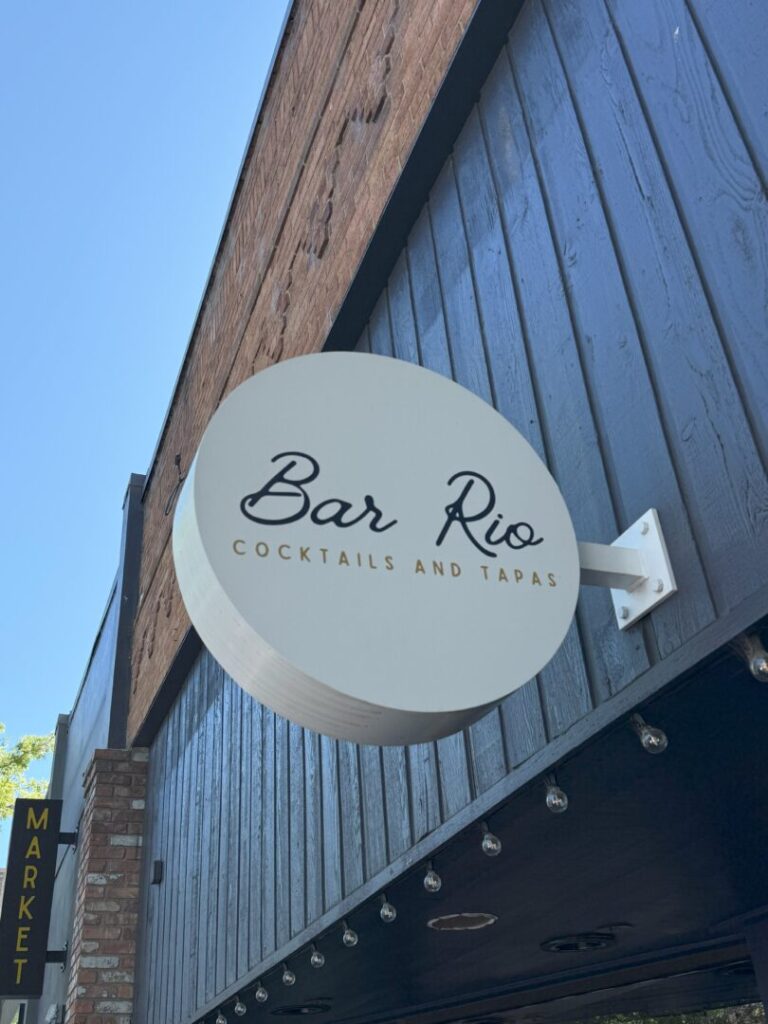 a Round White Sign with Black and Gold Text Reads bar Rio Cocktails and Tapas Mounted on a Blue Wood paneled Exterior This Spot Adds Charm to the Bend Restaurants Scene Strings of Lights Hang Above with a Partial Yellow market Sign on the Left