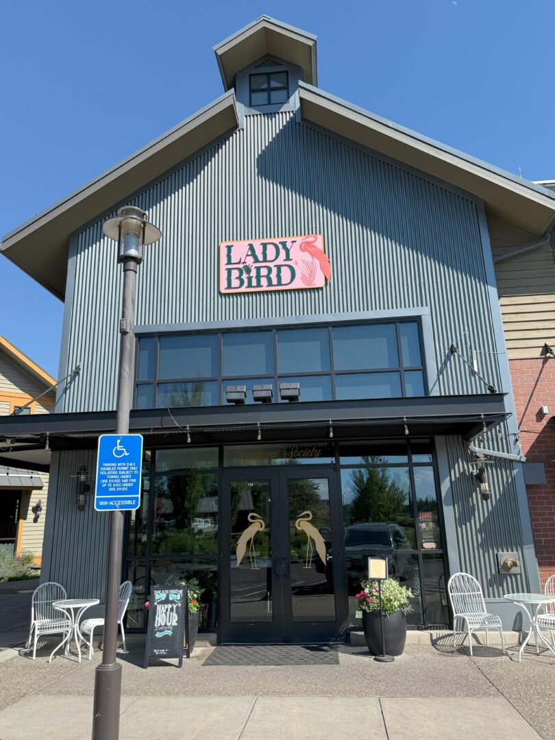 a Modern Gray Building Home to One of the Inviting Bend Restaurants Features a Sign Reading Lady Bird with a Bird Illustration Above Glass Doors Adorned with Bird Decals White Metal Tables and Potted Plants Flank the Entrance Near a Blue Handicap Sign