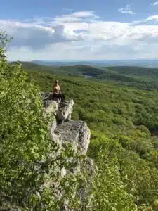 a Person Sits on the Edge of a Rocky Cliff Overlooking a Vast Green Forest One of the Best Hikes Around Frederick Md Trees Cover Rolling Hills to the Horizon with a Small Distant Lake Nestled in the Landscape Under a Partly Cloudy Sky