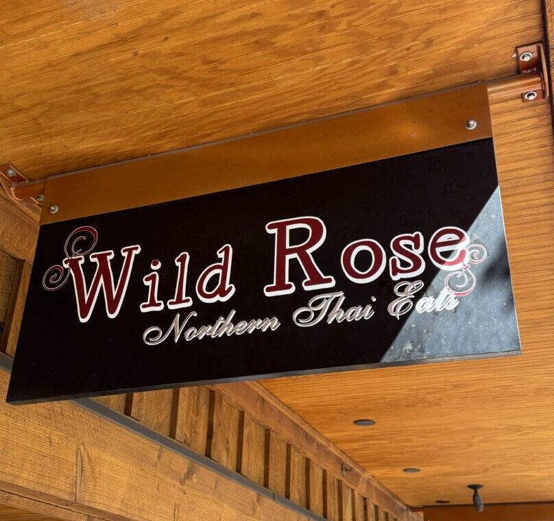 a Rectangular Black Sign with Wild Rose in Large White and Red Letters and Northern Thai Eats in Elegant White Script Below the Sign is Mounted Under a Wooden Ceiling with Visible Beams and Warm Lighting in Bend Restaurants