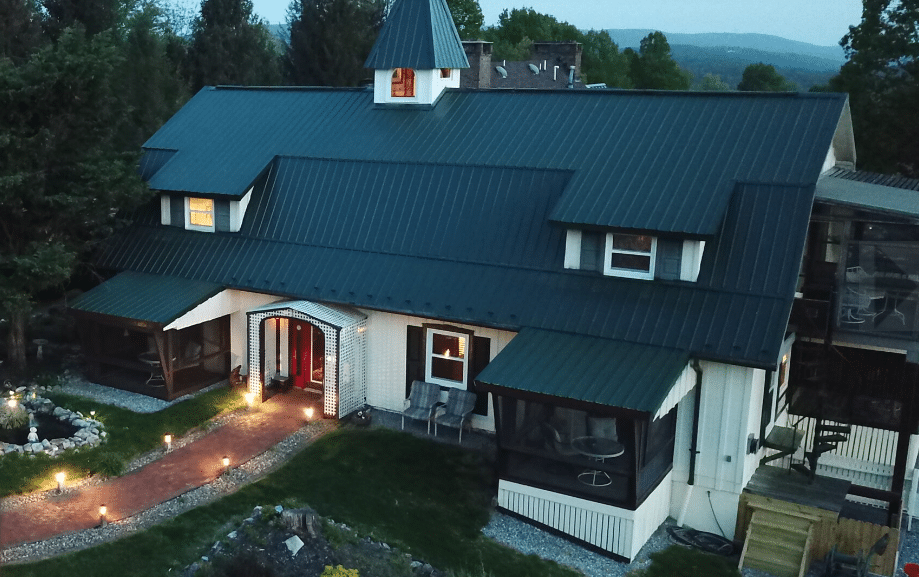 A large, dark-roofed house with dormer windows and a central peaked tower, resembling the charm of historic hotels in Frederick MD. It features screened porches, garden beds, a red-brick walkway, and green trees under a cloudy sky.