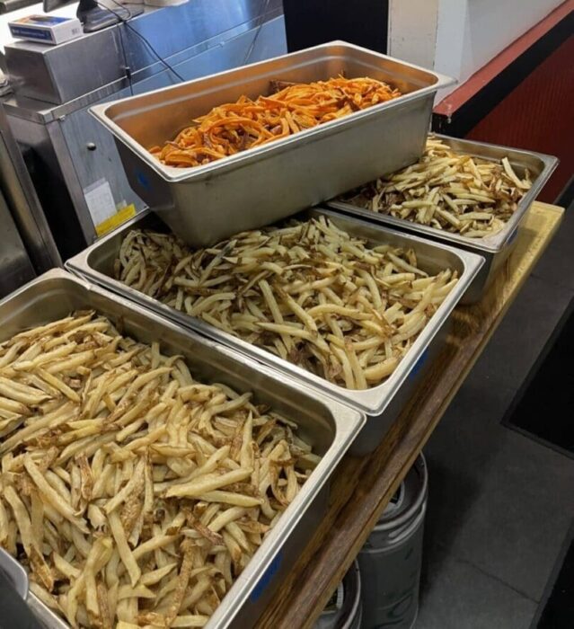 Four Metal Trays Filled with French Fries Are Stacked on a Counter in a Kitchen with Stainless Steel Appliancesa Classic Side to Enjoy with the Best Burger Frederick Md Has to Offer the Top Tray Features Orange Sweet Potato Fries Above Three Trays of Regular Fries