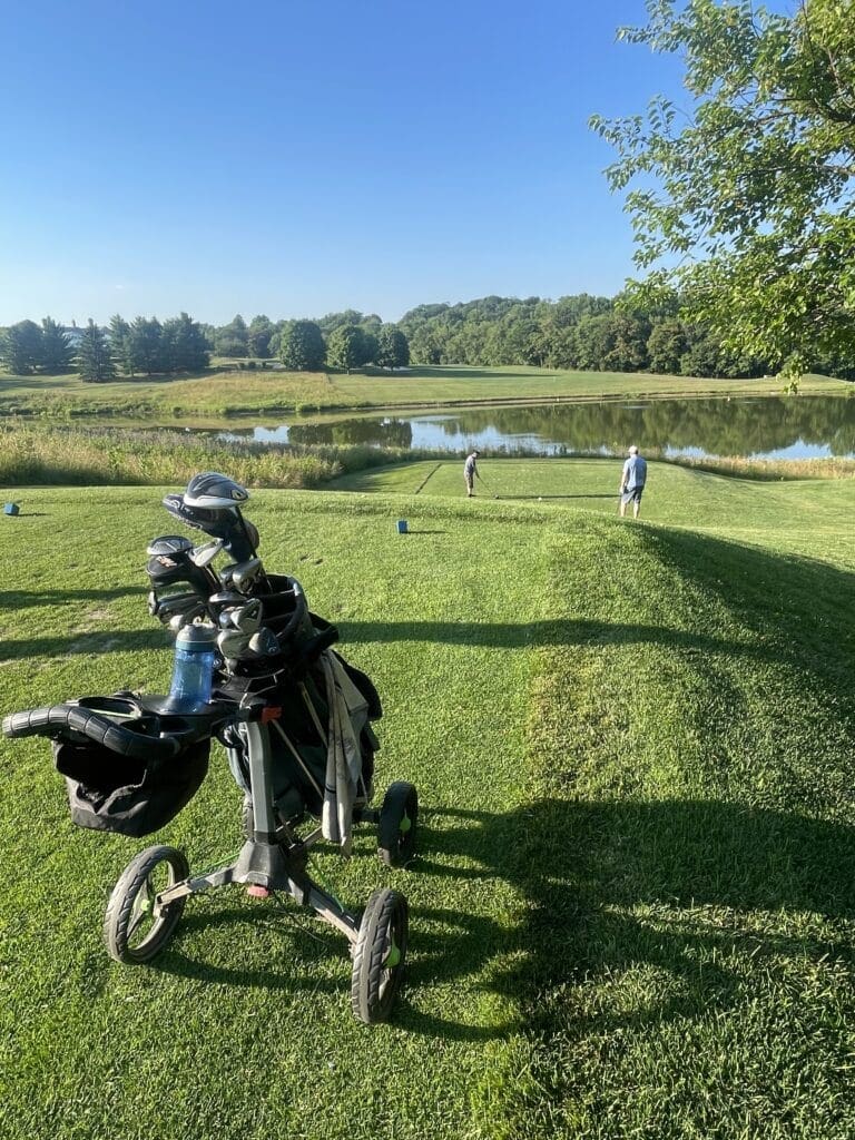 Rattlewood Hole 6 Tee Shot a Golf Bag on a Push Cart Sits on Green Grass in the Foreground Two Golfers Near a Tee by a Pond Surrounded by Trees and Under a Clear Blue Sky Enjoy a Sunny Day at the Mt Airy Md Course