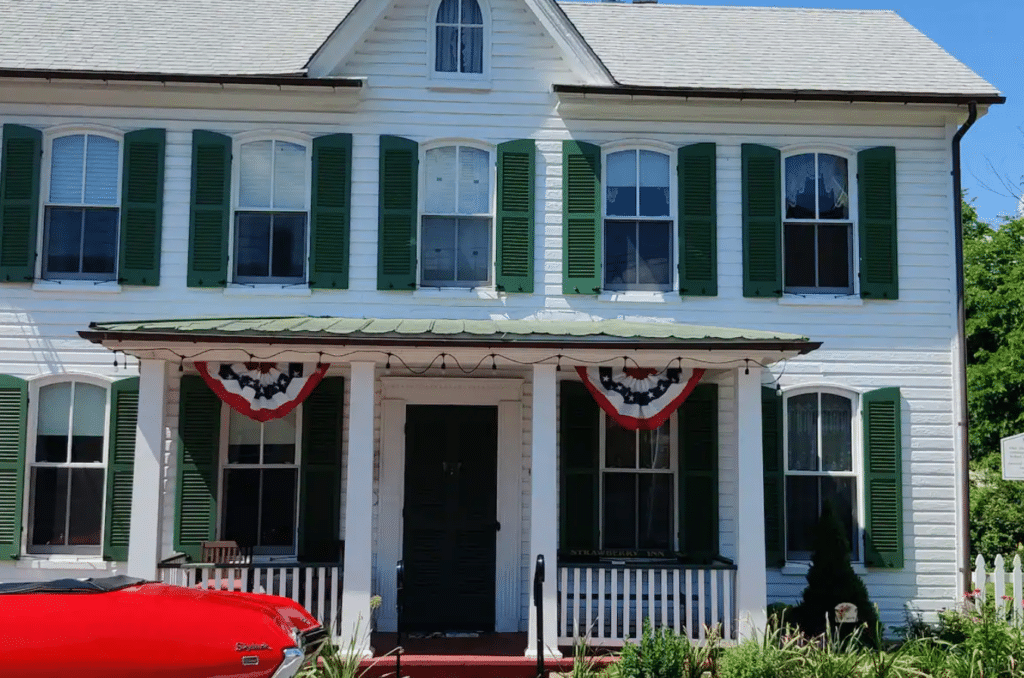 Strawberry Inn New Market a White Two Story House with Green Shutters a Metal Porch Roof and Patriotic Red White and Blue Bunting Above the Entrance Evokes Classic Charm in New Market Md a Red Car is Partially Visible by Shrubs and a White Picket Fence Lining the Porch