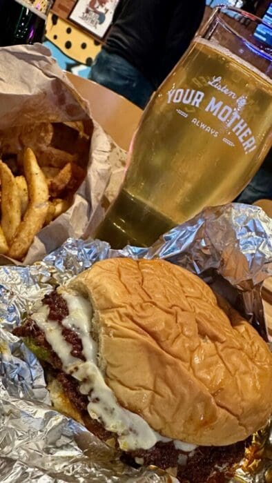 a Cheeseburger with a Toasted Bun and Melted Cheese Sits on Foil Next to Seasoned Fries Behind Them a Pint Glass of Golden Beer Reads Your Mothera Mouthwatering Scene from the Spot Known for the Best Burger Frederick Md Offers