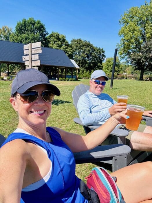 a Woman and Man Sit Outdoors in Adirondack Chairs at One of the Best Breweries in Frederick Md Smiling and Holding Up Plastic Cups of Beer Both Wear Sunglasses Hats and Casual Summer Clothes Framed by Green Grass Trees and a Pavilion Under Clear Blue Skies