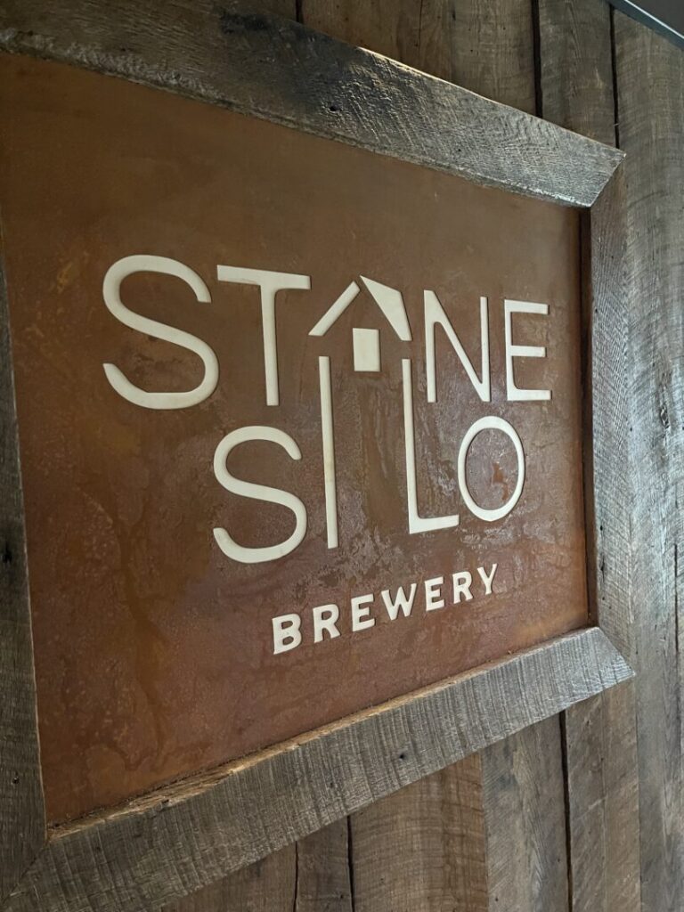 Stone Silo Near Frederick Md a Rustic Wooden Sign Displays the Text Stone Silo Brewery in White with the a in Stone Styled As a Roof with a Small Chimney to Resemble a House Set Against a Weathered Rusty Orange Background and Rough Wooden Frame It Captures New Market Md Charm