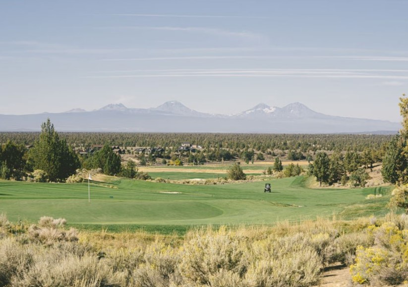 Brasada canyons golf course bend a Scenic Golf Course with Lush Green Fairways and a Golf Cart Sits in the Center Sagebrush and Trees Surround the Course Near Rolling Forests and Snow capped Mountainsperfect for Those Seeking Where to Stay in Bend Oregon