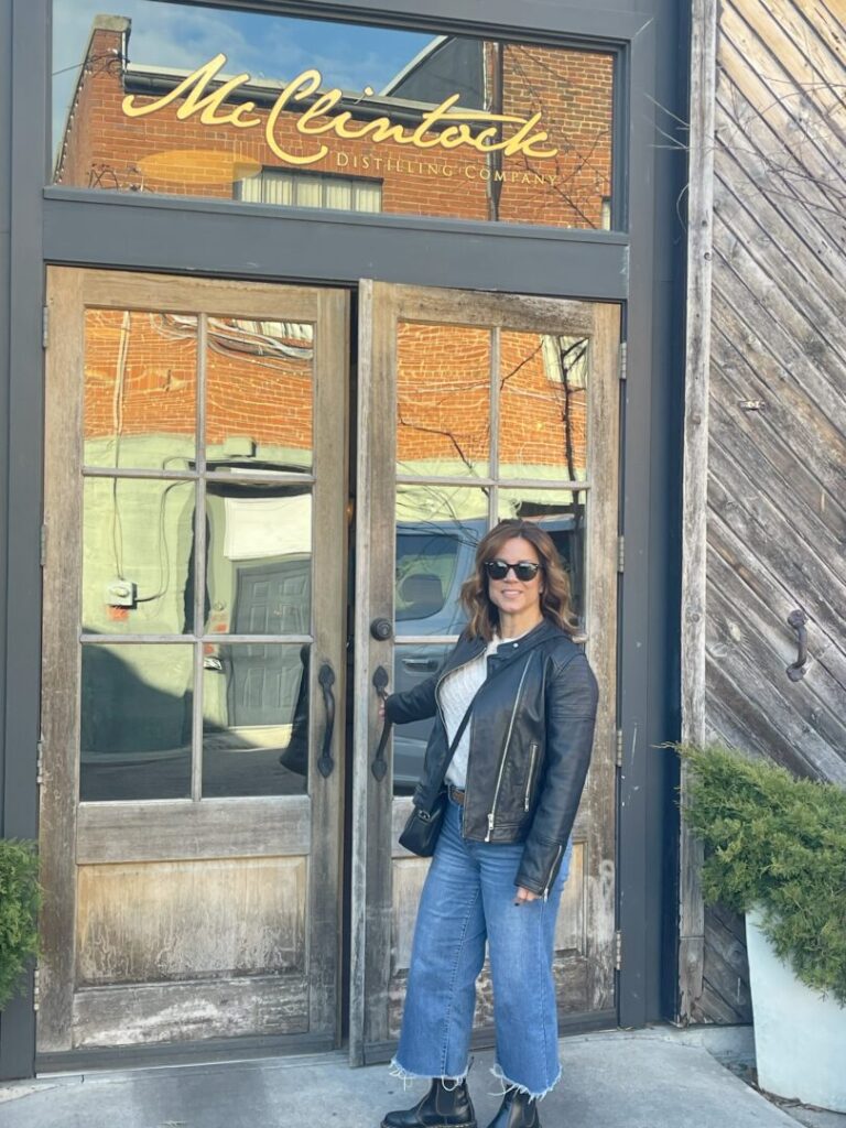 Nicole Mcclintocks Entrance a Woman Wearing Sunglasses a Black Leather Jacket Gray Shirt Blue Wide Leg Jeans and Black Boots Stands Smiling in Front of Wooden Double Doors with a Gold Mcclintock Distilling Company Sign Abovean Iconic Spot Near Top Hotels in Frederick Md Green Shrubs in Planters Flank the Entrance