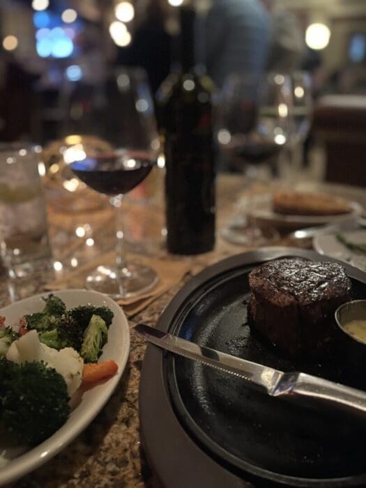 a Dimly Lit Restaurant Table at the Best Steakhouse Frederick Md Features a Steak on a Black Plate a Knife Sauce Steamed Vegetables Red Wine Glasses a Bottle of Wine and Sparkling Water with Warm Lights in the Background