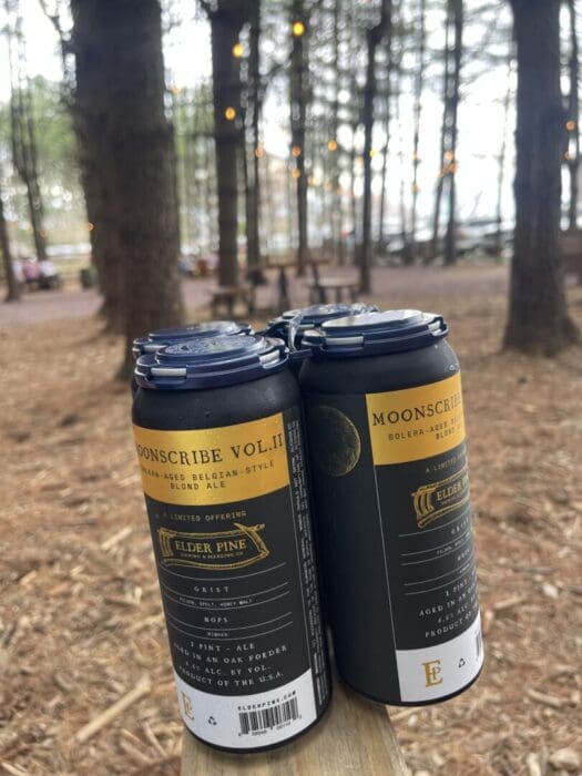 Four Black and Gold Cans of Moonscribe Vol Ii Elder Pine Ale from One of the Best Breweries in Frederick Md Sit on a Wooden Surface Outdoors Surrounded by Tall Trees and Warm String Lights for a Cozy Forest Vibe with Blurred Benches and Tables