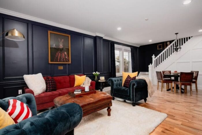 a Stylish Living Room Inspired by Boutique Hotels in Downtown Frederick Md with Dark Navy Paneled Walls a Red Sofa Green Velvet Chairs and a Whimsical Rabbit Portrait Above Hardwood Floors Lead to a Dining Area and White Staircase in the Background