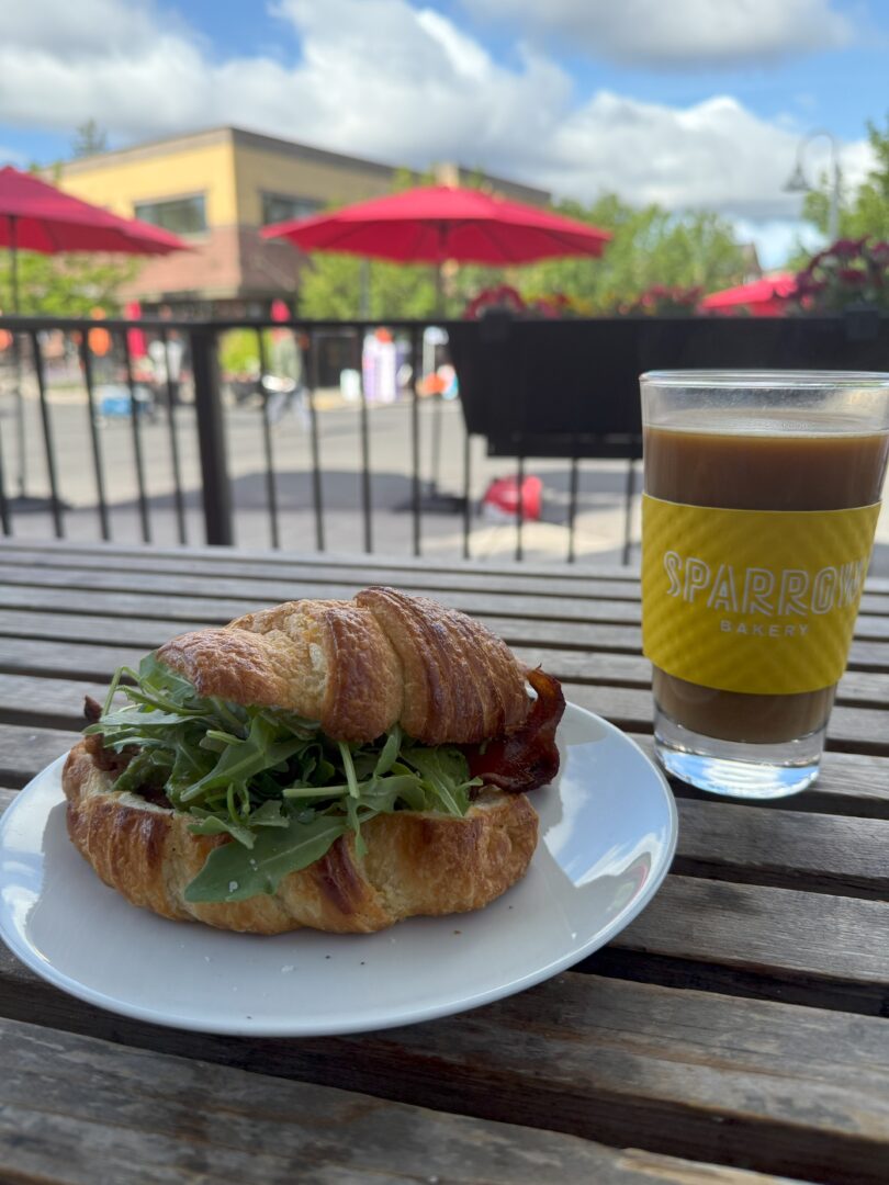 Bacon Breakfast Sandwich Sparrow Bakerty a Croissant Sandwich Filled with Arugula Sits on a White Plate Next to a Glass of Iced Coffee Labeled Sparrow Bakery Atop a Wooden Table Outside Red Umbrellas Greenery and Buildings Are Visible in the Sunny Background