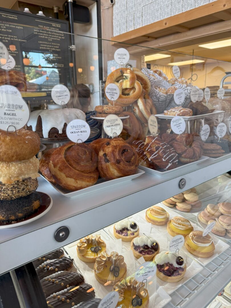 Baked Goods Sparrow Bakery a Bakery Display Case Filled with Pastries Including Stacked Donuts Cinnamon Rolls Braided Breads and Croissants on the Top Shelf with Tarts and Cakes Topped with Fruit and Whipped Cream on the Lower Shelf Labels Mark Each Item