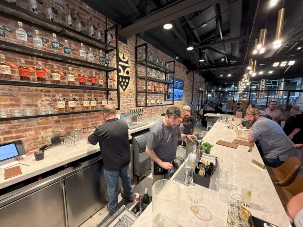 a Busy Bar with Bartenders Preparing Drinks Behind a Marble Counter Liquor Bottles Are Neatly Arranged on Wall Shelves Above Among the Best Bars in Frederick It Boasts Modern Exposed Brick Walls Pendant Lighting and a Lively Atmosphere