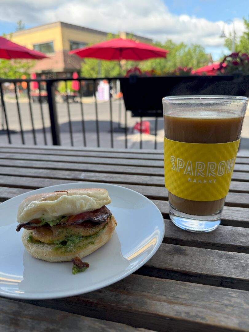 Breakfast Sparrow Bakery a Breakfast Sandwich with Bacon Egg and Green Arugula Sauce Sits on a White Plate Next to a Glass of Iced Coffee Labeled Sparrow Bakery on a Wooden Outdoor Table the Background Shows Red Umbrellas Trees and a Building Under a Partly Cloudy Sky