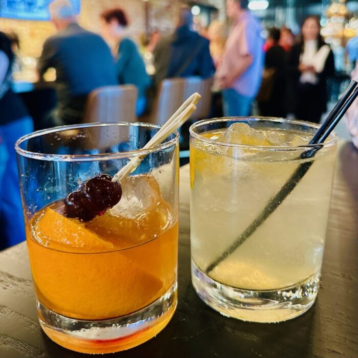 Craft Cocktails True Standard Distilling Two Cocktails in Short Glasses Sit on a Black Table at a Lively Spot Near a Coffee Shop in Frederick Md One Drink Features an Orange Slice and Cherries the Other is Pale Yellow with Ice and a Black Straw Blurred People and a Busy Bar Fill the Background
