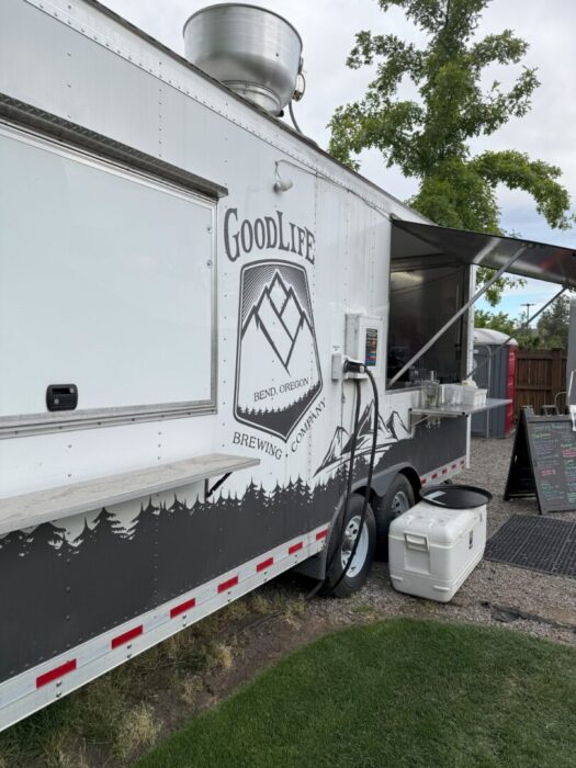 a White Food Truck Labeled Goodlife Brewing Company Bend Oregon Features a Mountain Logo and Tree Silhouettes the Side Window is Open with Hoses and Equipment Attached a White Cooler Sits Nearby and a Menu Board is Visible in the Background a White Food Truck Labeled Goodlife Brewing Company Bend Oregon Features a Mountain Logo and Tree Silhouettes the Side Window is Open with Hoses and Equipment Attached a White Cooler Sits Nearby and a Menu Board is Visible in the Background