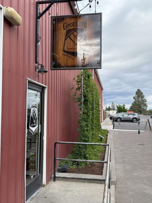 a Red Metal Building with a Glass Door and a Wooden Sign Reading Goodlife Brewing Company Green Climbing Plants Grow Up the Side and the Sidewalk Leads to a Mostly Empty Parking Lot Under a Cloudy Sky a Red Metal Building with a Glass Door and a Wooden Sign Reading Goodlife Brewing Company Green Climbing Plants Grow Up the Side and the Sidewalk Leads to a Mostly Empty Parking Lot Under a Cloudy Sky
