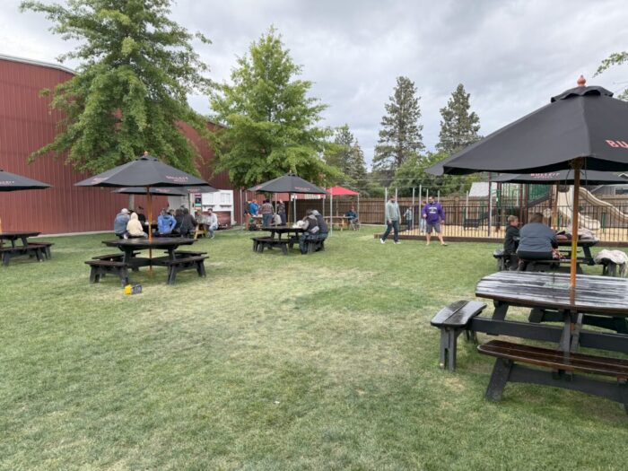 People Sit at Round Black Picnic Tables with Umbrellas on a Grassy Lawn Some Children and Adults Stand and Walk Near a Playground Behind a Fence Trees and a Red Building Are Visible in the Background Under a Cloudy Sky People Sit at Round Black Picnic Tables with Umbrellas on a Grassy Lawn Some Children and Adults Stand and Walk Near a Playground Behind a Fence Trees and a Red Building Are Visible in the Background Under a Cloudy Sky