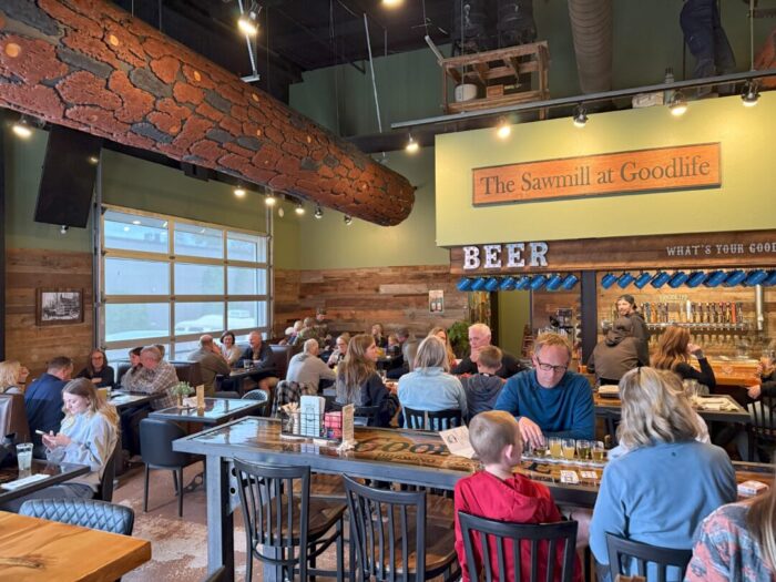 a Lively Restaurant Interior with Wooden Decor and a Faux Log Beam Diners Sit at Tables Chatting and Eating a Sign Reads the Sawmill at Goodlife Above a Bar Area Displaying Beer the Atmosphere is Warm and Social with Families and Groups Gathered a Lively Restaurant Interior with Wooden Decor and a Faux Log Beam Diners Sit at Tables Chatting and Eating a Sign Reads the Sawmill at Goodlife Above a Bar Area Displaying Beer the Atmosphere is Warm and Social with Families and Groups Gathered