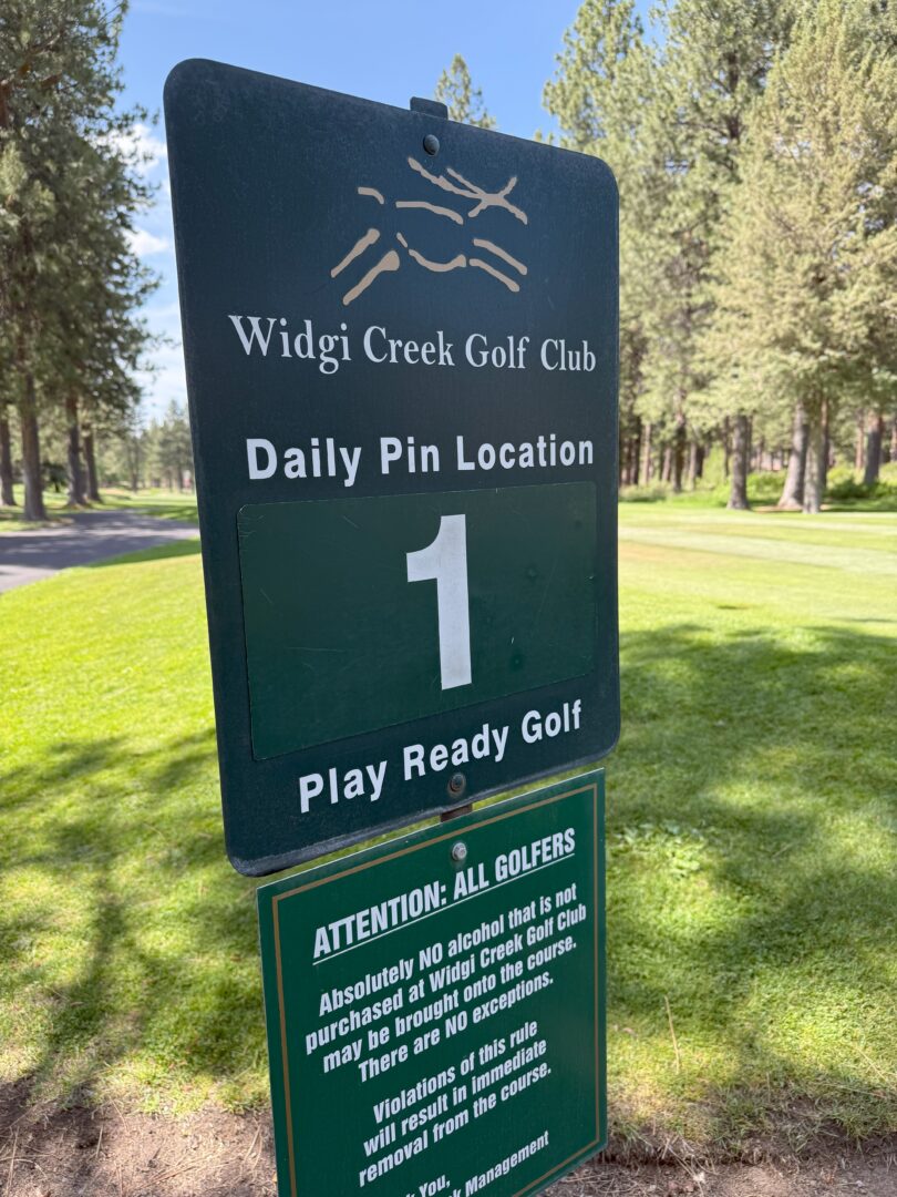 a Green Sign at Widgi Creek Golf Club Displays Daily Pin Location 1 and Play Ready Golf Below a Notice Warns That Outside Alcohol is Not Allowed and Violations May Result in Removal Trees and Green Grass Are Visible in the Background a Green Sign at Widgi Creek Golf Club Displays Daily Pin Location 1 and Play Ready Golf Below a Notice Warns That Outside Alcohol is Not Allowed and Violations May Result in Removal Trees and Green Grass Are Visible in the Background