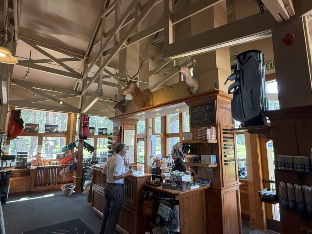 a Man Stands at a Wooden Counter in a Golf Pro Shop with Large Windows Behind the Counter a Woman Assists Him Golf Gear Apparel and Displays Fill the Shop Two Deer Mounts Are on the Beam Above and Sunlight Streams Through the Tall Windows a Man Stands at a Wooden Counter in a Golf Pro Shop with Large Windows Behind the Counter a Woman Assists Him Golf Gear Apparel and Displays Fill the Shop Two Deer Mounts Are on the Beam Above and Sunlight Streams Through the Tall Windows