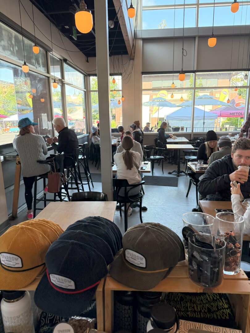 Sparrow Bakery Bend or a Bright Modern Café with Large Windows and Hanging Lights Where People Sit at Tables and Bar Seating Chatting or Working in the Foreground Hats Cups and Coffee Beans Are Displayed on a Wooden Counter Outdoor Seating and Umbrellas Are Visible Outside