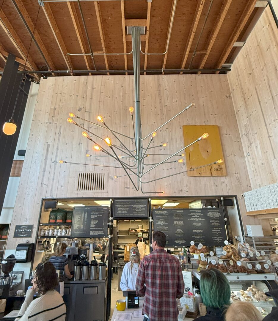 Sparrow Bakery Bend Oregon a Modern Cafe Interior with Light Wood Walls an Industrial Chandelier with Exposed Bulbs and a High Unfinished Ceiling Customers Stand at the Counter Baked Goods and Menus Are Visible a Yellow Abstract Painting Hangs on the Back Wall