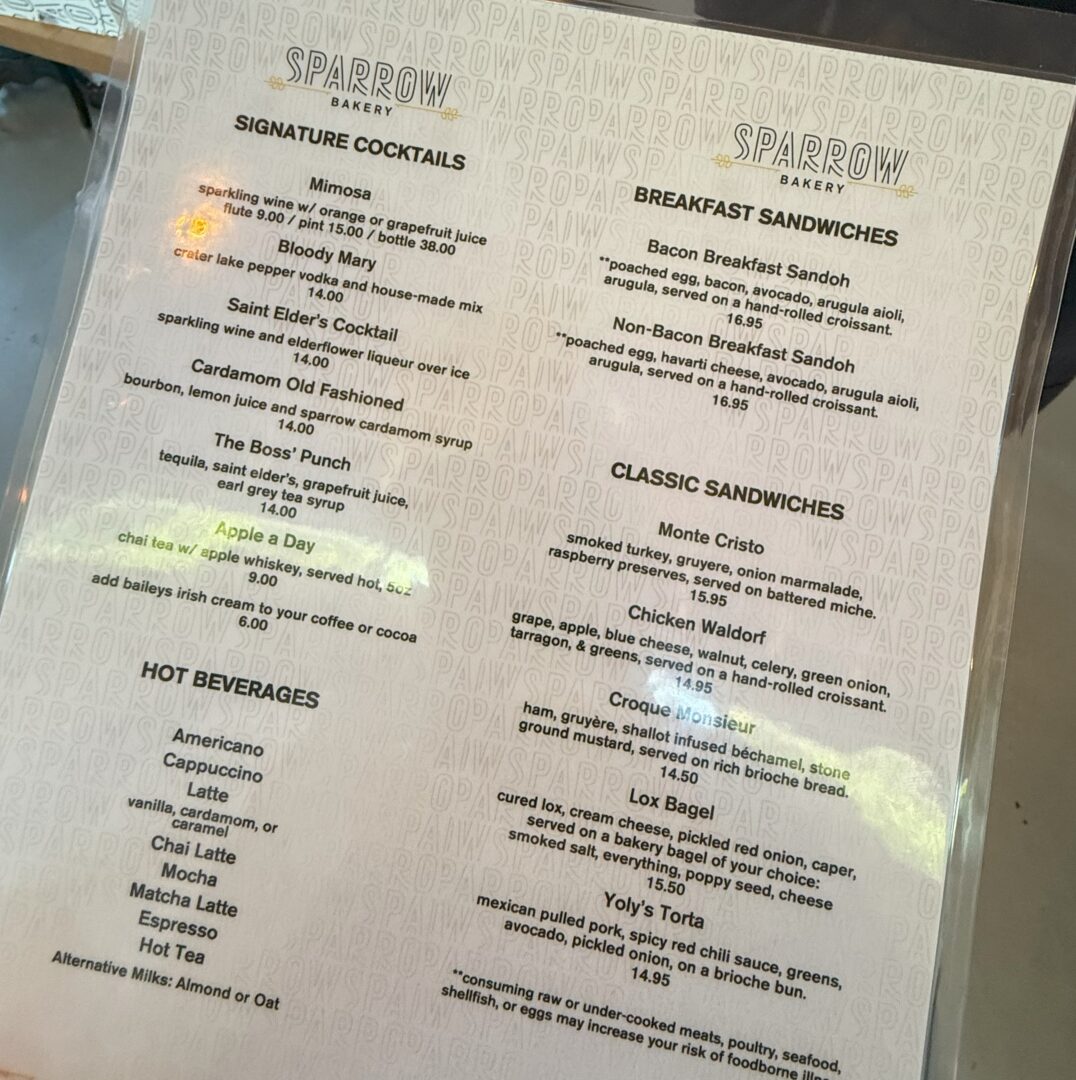 Sparrow Bakery Breakfast Menu a Laminated Café Menu on a Table Lists Signature Cocktails Hot Beverages Breakfast Sandwiches and Classic Sandwiches Items Include Mimosas Bloody Mary Café Drinks Bacon and Egg Sandwiches Monte Cristo Chicken Waldorf Croque Monsieur and Lox Bagel
