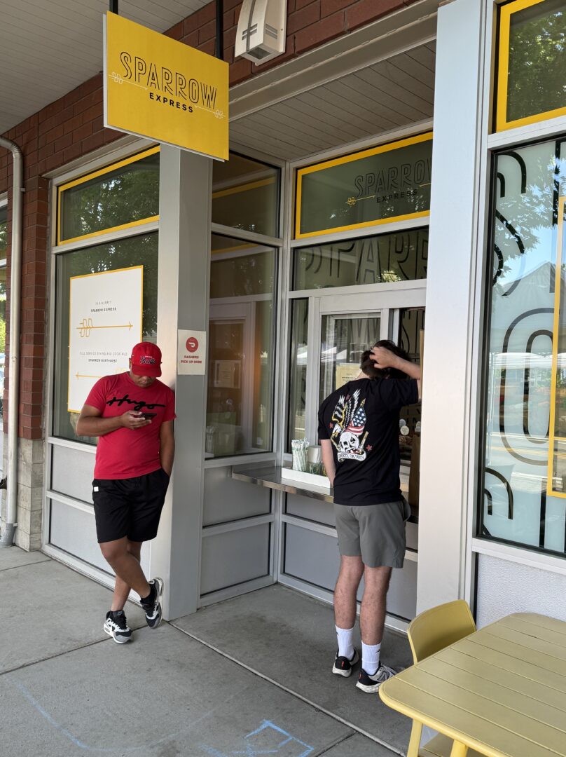 Sparrow Bakery Express Counter Two People Wait Outside Sparrow Bakery One in a Red Hat and Shorts Looks at His Phone and Leans Against the Wall the Other in a Black Shirt with a Colorful Back Design Stands by the Order Window Yellow Chairs and Tables Are Nearbyperfect for an Ocean Roll Break