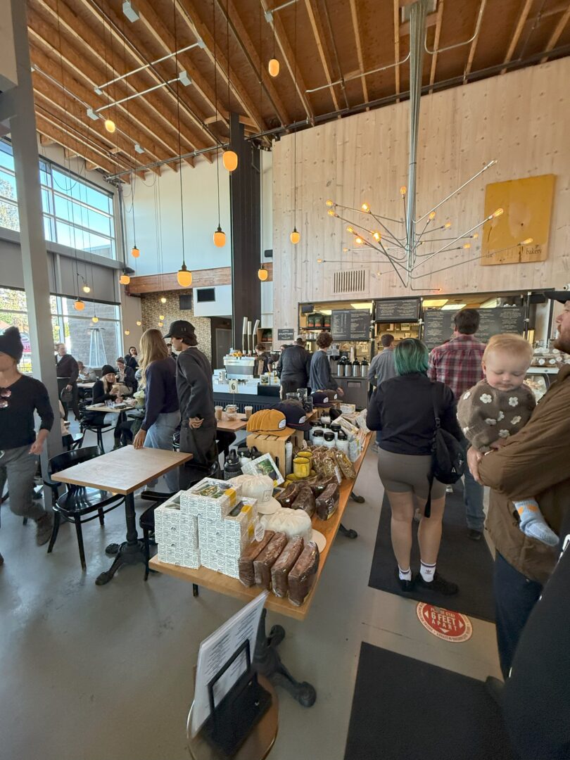 Sparrow Bakery Interior a Busy Modern Café with High Ceilings Hanging Lights and Large Windows People Wait in Line at the Counter and Sit at Tables Breads and Packaged Goods Are Displayed on a Central Table a Man Holds a Baby in the Foreground Bright Lively Atmosphere