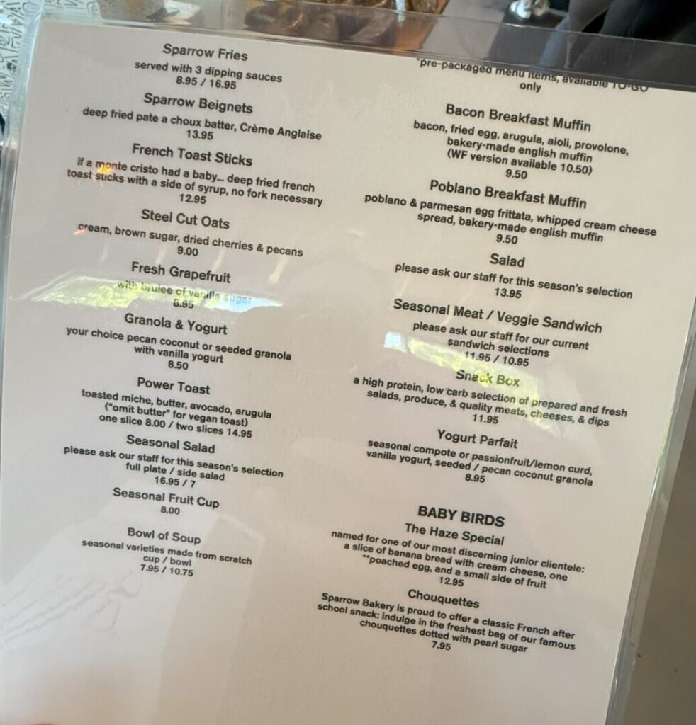 Sparrow Bakery Menu a Printed Cafe Menu on a White Background Lists Breakfast and Brunch Items Such As Sparrow Fries French Toast Sticks Steel Cut Oats Granola Toast Options Muffins Sandwiches Salad Yogurt Parfait Soups and Kids Meals Reflections Are Visible on the Menu